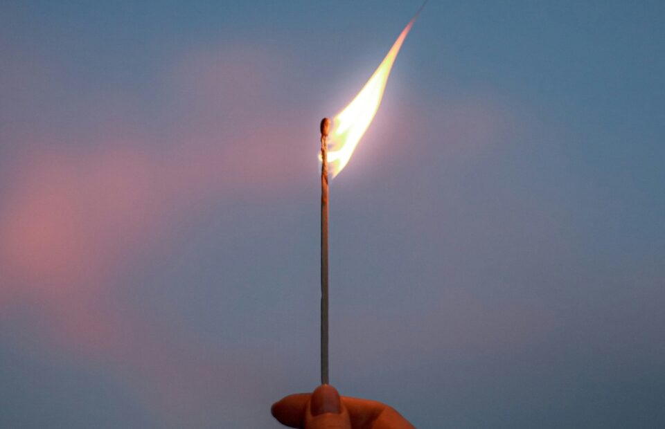 A hand holds a lit match with a bright flame against a twilight sky. The background is mostly blue with soft pinkish clouds.