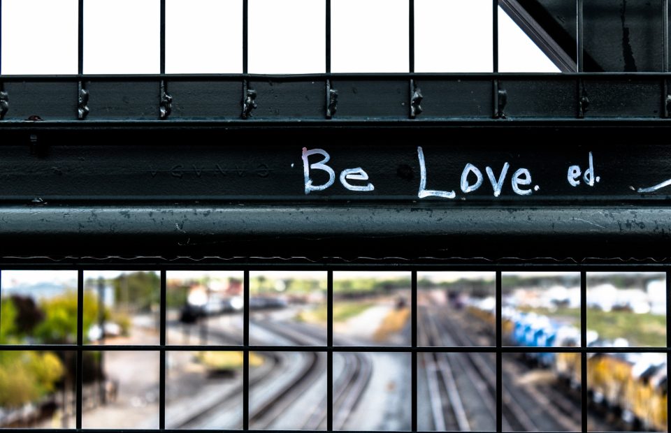 A close-up of a metal fence with graffiti reading "Be Love." Overhead train tracks and a blurred parking lot are visible in the background.