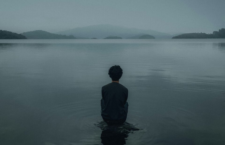 A person in a suit stands waist-deep in a calm, expansive lake, facing away toward distant hills and a moody, overcast sky.