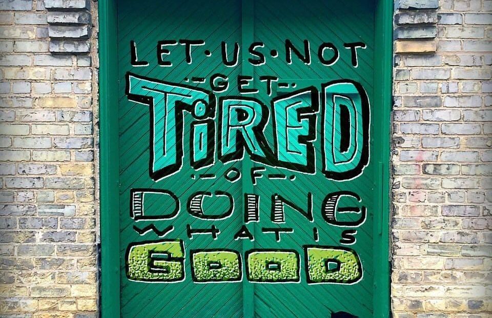 A green door with a motivational graffiti message: "Let us not get tired of doing what is good," referencing Galations 6:9, with a brick wall background.