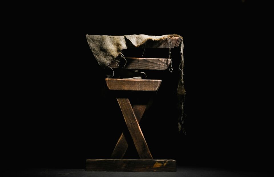 A wooden chair with a tattered cloth draped over the top, set against a black background with dramatic lighting highlighting its texture and details.
