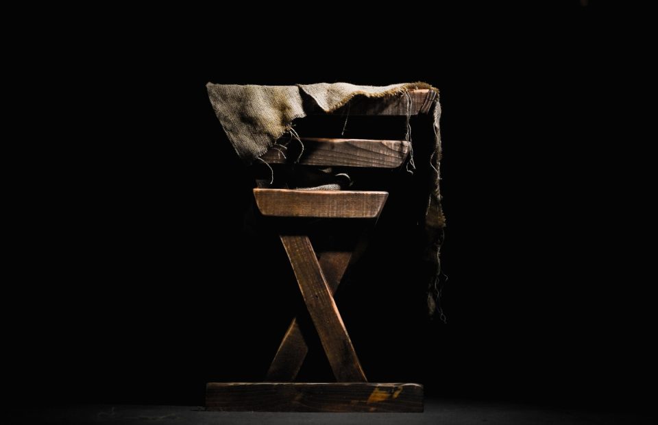 A worn wooden chair with a frayed, distressed piece of fabric draped over the top, set against a black background with light highlighting the chair's texture.