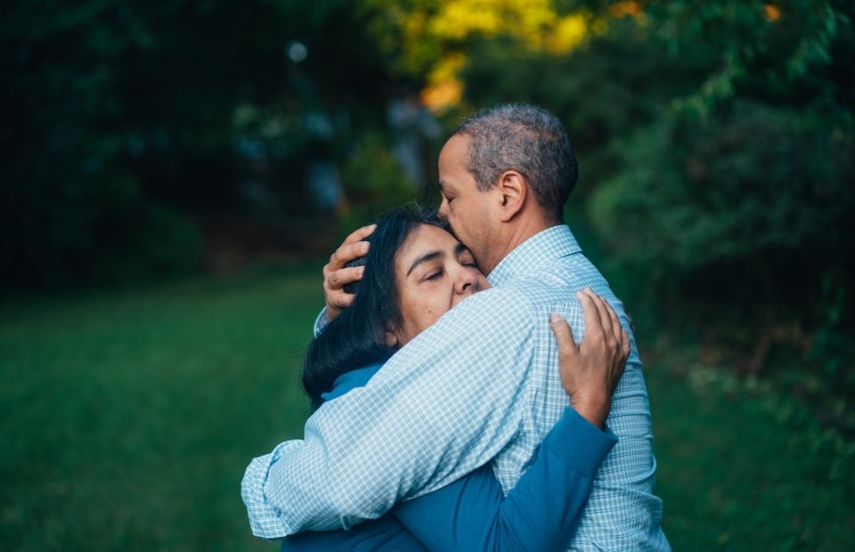 A man and woman embrace outdoors with trees in the background; the man kisses the woman on the forehead as she closes her eyes and holds on to him.