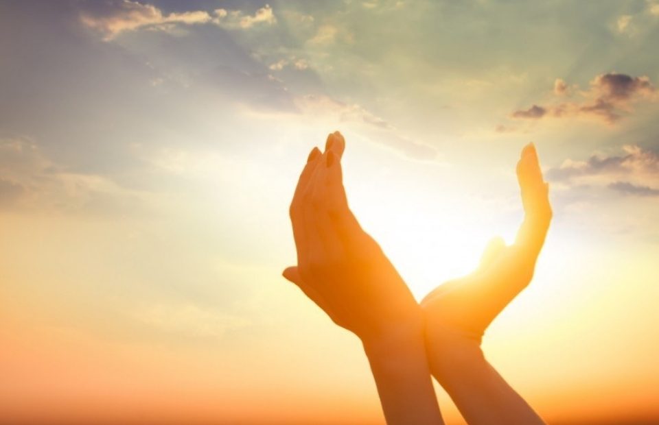 Two hands reaching towards the sky during sunset, with the sun partially obscured by the hands, casting a warm glow. The sky has scattered clouds and vibrant colors.