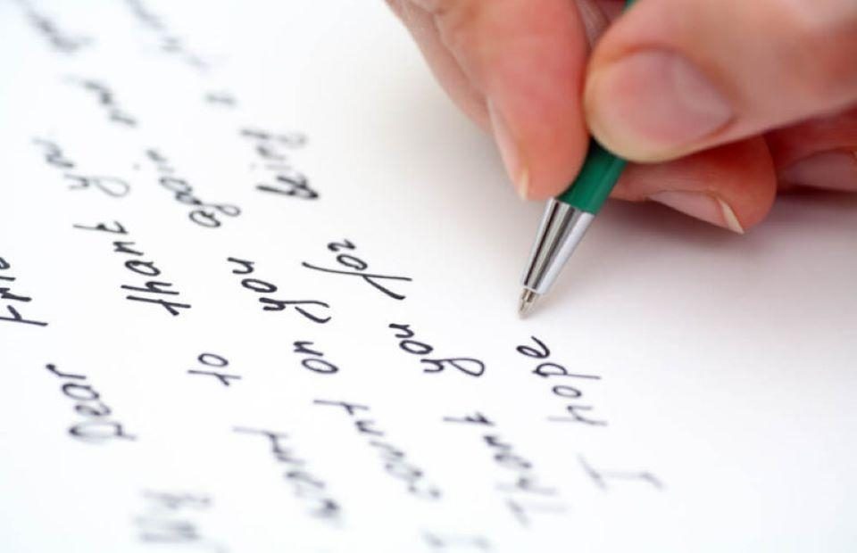 Hand writing a letter with a green pen on white paper.