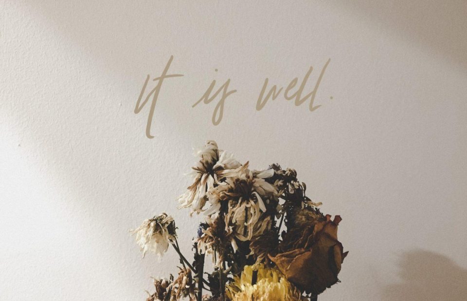 Dried, wilted flowers arranged in front of a light-colored wall with the words "it is well" written in cursive above.