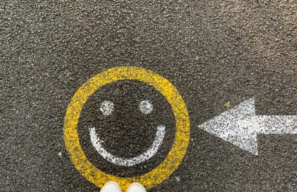 Person standing on pavement next to a smiley face painted with a yellow circle and white features, and an arrow pointing right.