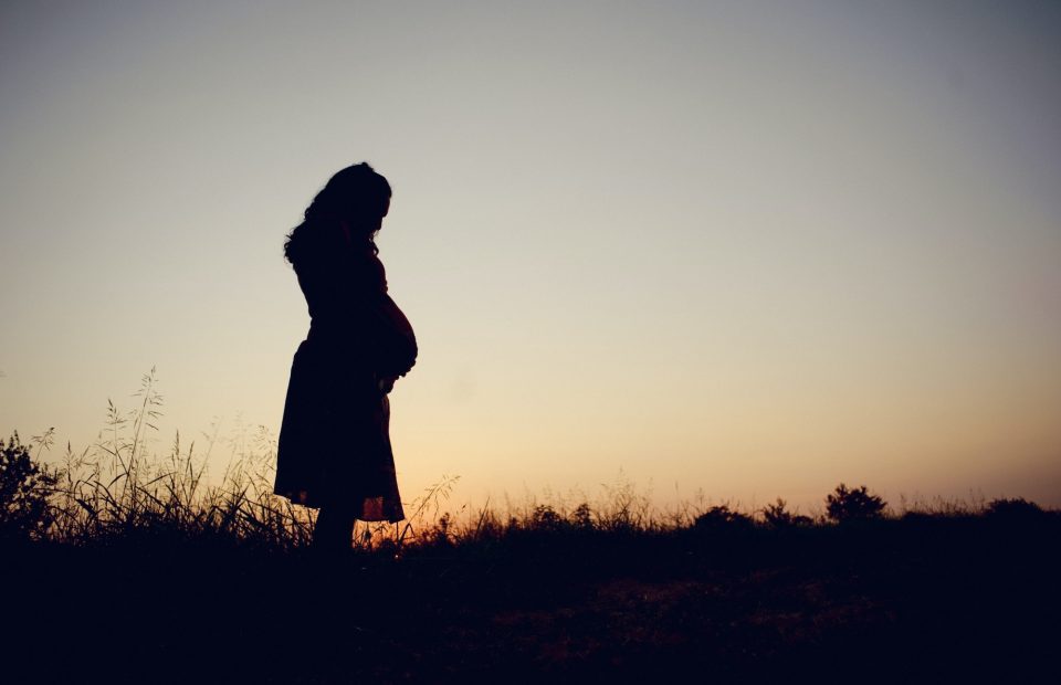 A pregnant woman stands outdoors on a hill during sunset, holding her belly and looking down contemplatively.