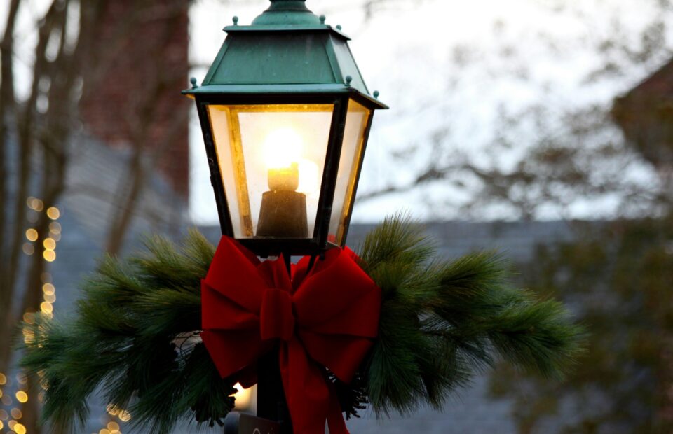 A decorated outdoor lantern with a red bow and pine branches, illuminated against a blurred background of trees and festive lights.