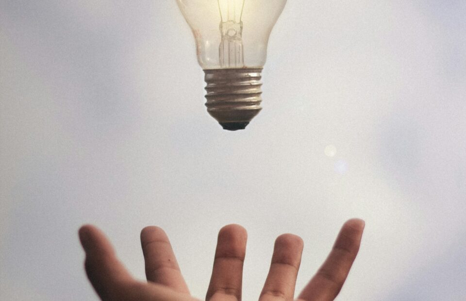 A hand appears to be tossing a lit lightbulb into the air against a cloudy sky background.