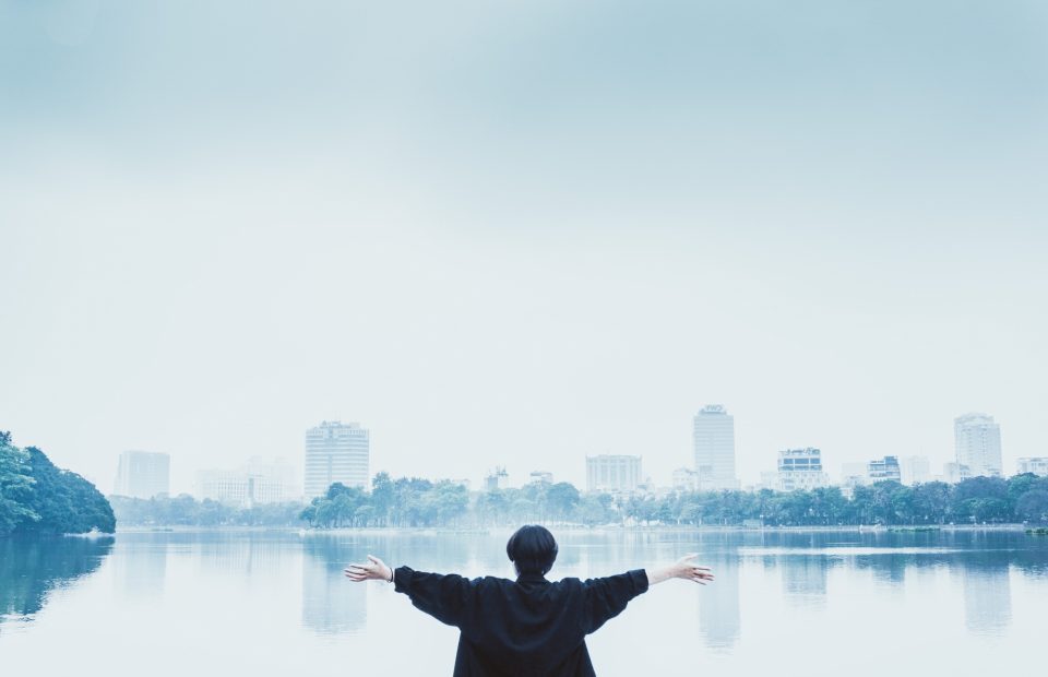 A person standing by a lake with arms outstretched, facing a city skyline in the distance, under a clear, light blue sky.