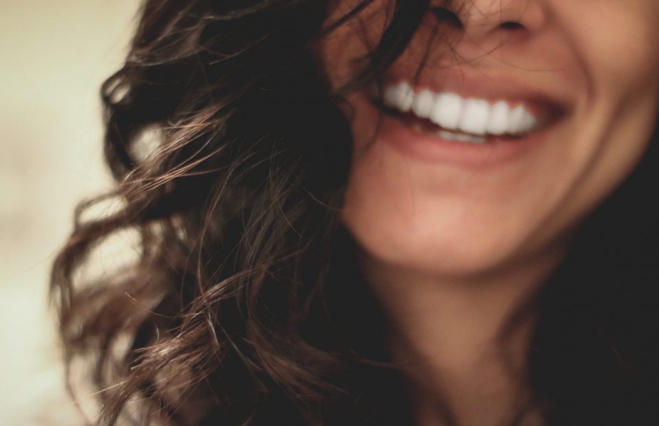 Close-up of a woman smiling, showing white teeth, with dark wavy hair partially covering her face.