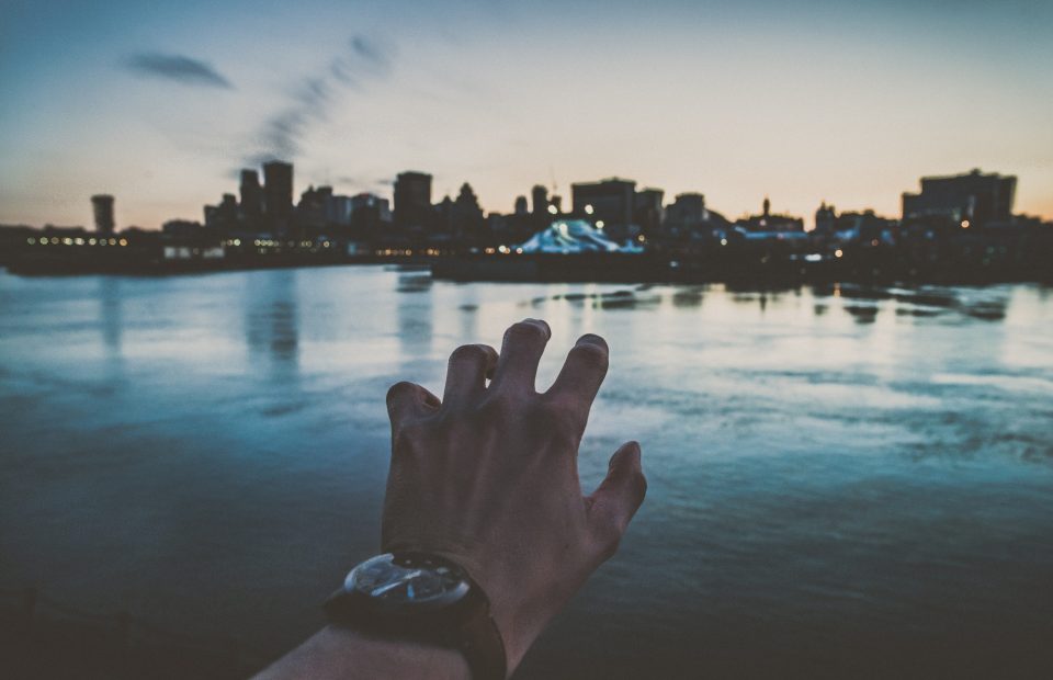 A hand reaching towards a city skyline across a body of water during sunset or twilight, with the silhouette of buildings and a church spire visible.