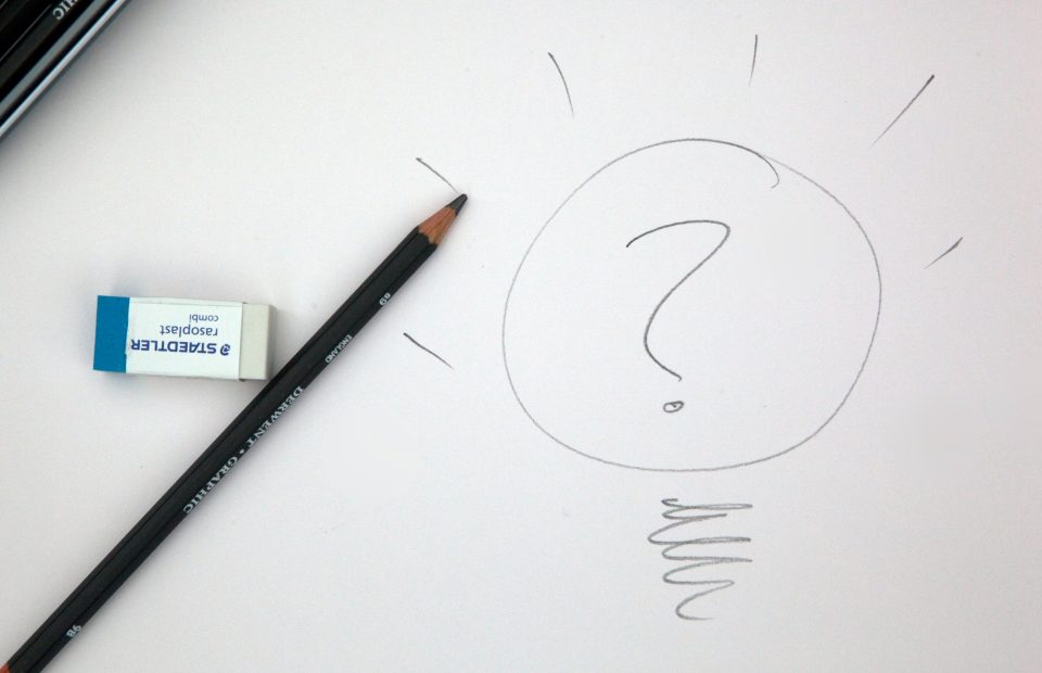 A pencil with an eraser, a paper with a hand-drawn lightbulb and a question mark inside it, and a small eraser on a white surface.