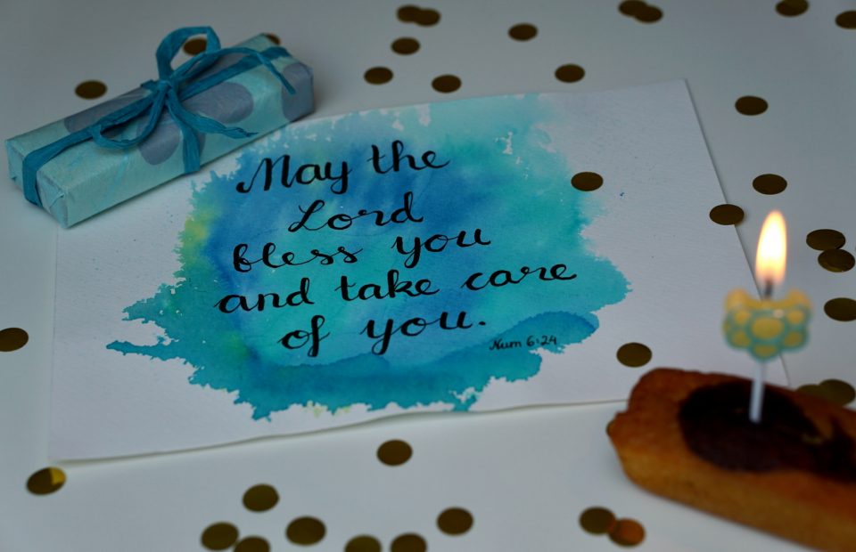 A greeting card with a blue watercolor background displays the message, "May the Lord bless you and take care of you," with a lit birthday candle, a wrapped gift, and scattered gold confetti on a white surface.