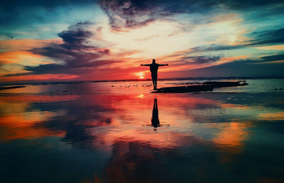 A person stands with arms outstretched on a rocky shoreline during a vibrant sunset, with colorful clouds and their reflection on calm water.