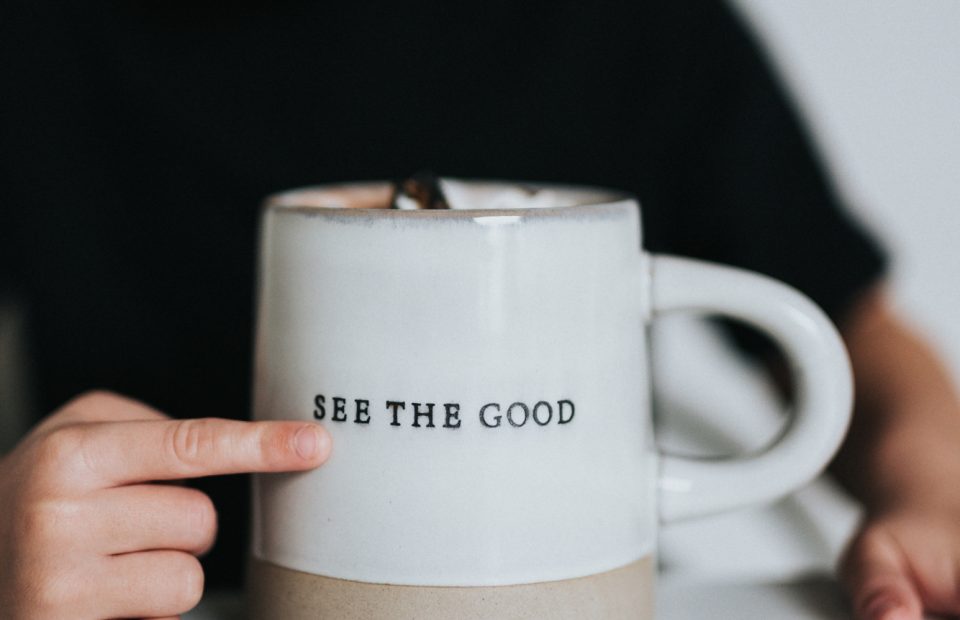 A child with a blurred face points to a white mug with "SEE THE GOOD" written on it, sitting on a white surface against a plain background.