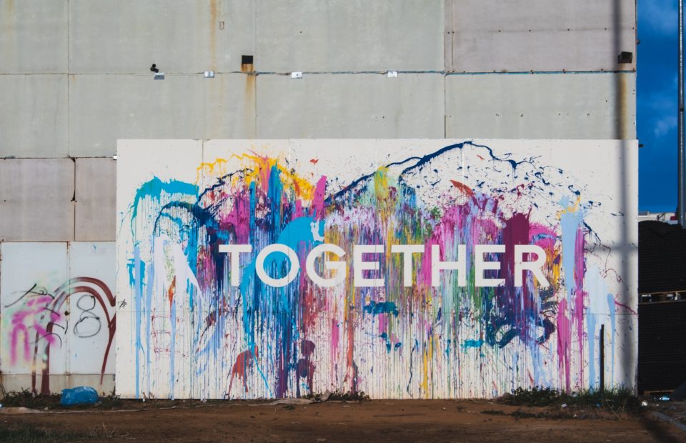 A colorful splatter-painted wall with the word "TOGETHER" in white letters, against a background of a concrete building wall and a partly cloudy blue sky.