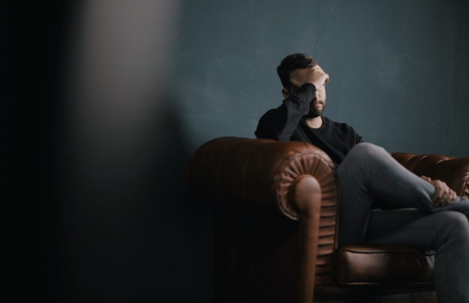 A man with a beard sitting on a brown leather couch, holding his forehead with his right hand, appears distressed or upset in a dimly lit room.