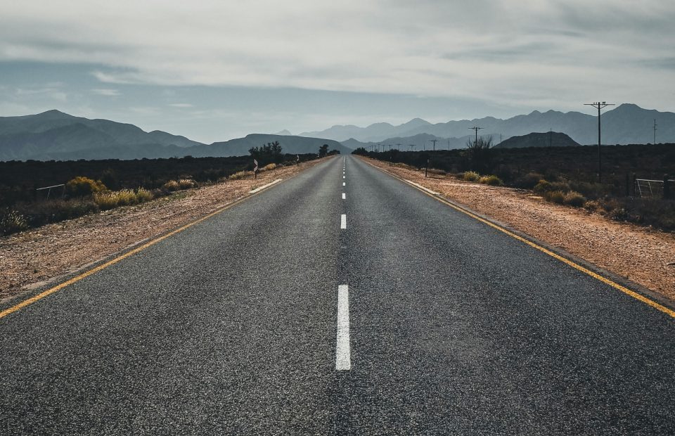 A straight, empty asphalt road stretches into the distance with mountains in the background and cloudy sky overhead. The road is flanked by desert terrain.