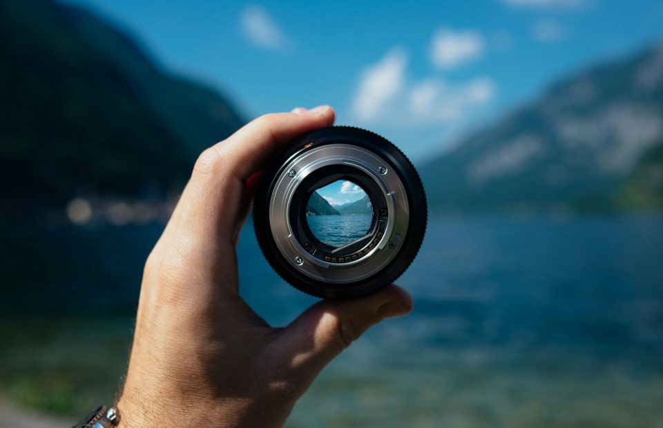A hand holds a camera lens, capturing a scenic view of a lake with mountains and clouds in the background, seen through the lens.