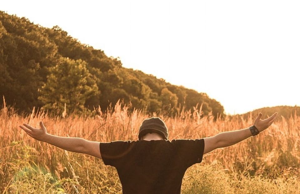 A person in a Pizza Ranch uniform stands in a field during sunset with arms outstretched, facing away from the camera, surrounded by tall grass and hills.