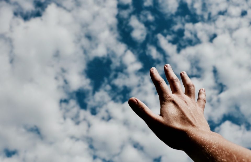 A hand reaching up towards a cloudy sky, with the arm partially visible and the sky filled with fluffy white clouds.