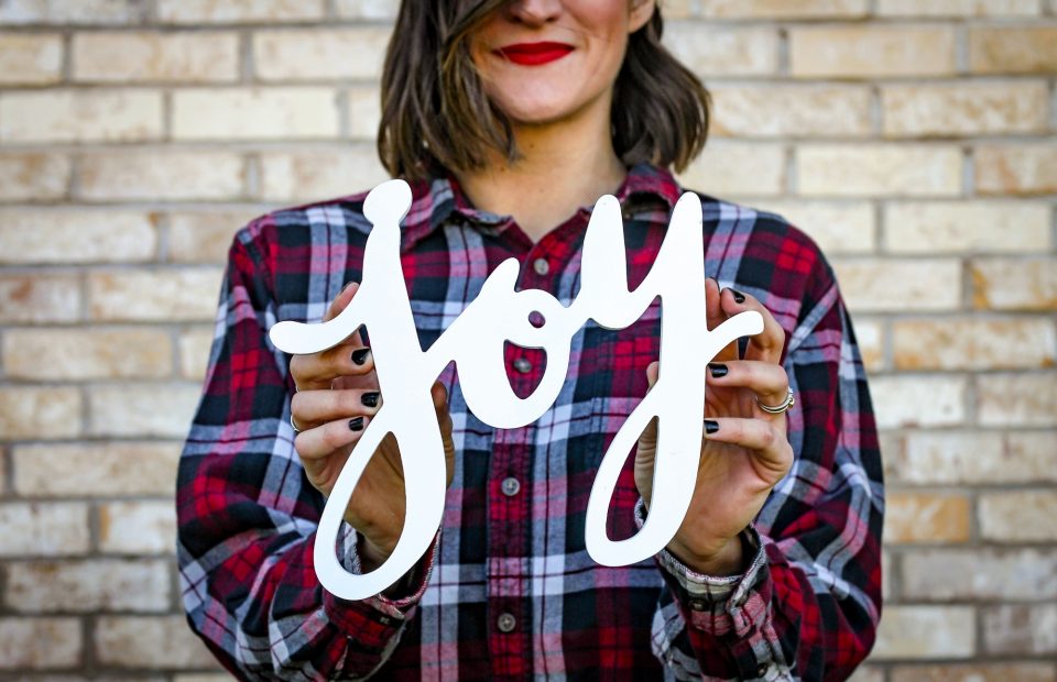 A woman with shoulder-length dark hair, red lipstick, wearing a red and black plaid shirt, holds a white "joy" sign in front of a brick wall.