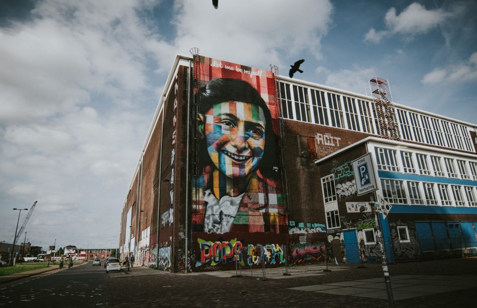 A tall building with colorful graffiti and a large portrait of a smiling man on the side, with birds flying above and people walking nearby.