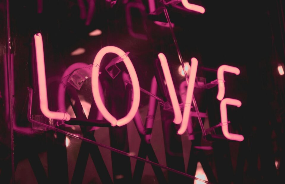 A neon pink sign spelling "LOVE" mounted on a reflective surface, glowing in a dark environment.