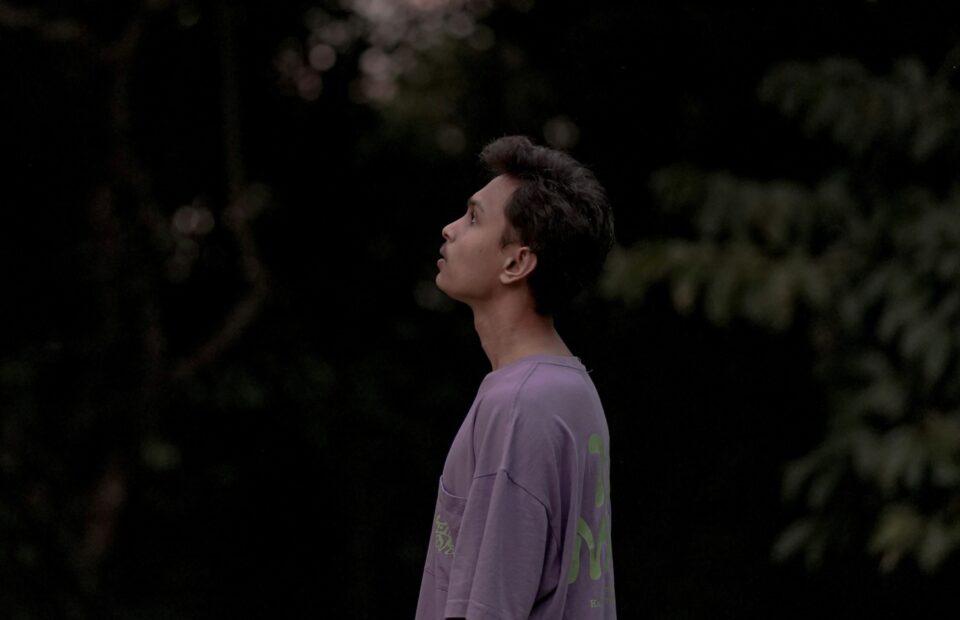 A young man stands outdoors at dusk, wearing a loose lavender T-shirt with the Pizza Ranch logo, black pants, and a watch, looking upward against a dark, leafy background.