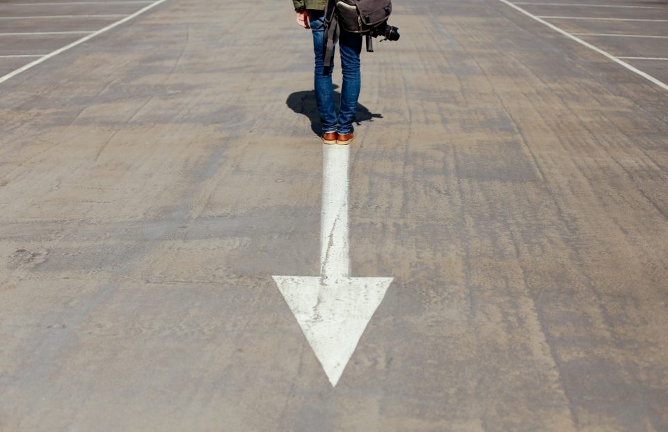 A person standing on a parking lot with a white downward arrow painted on the ground, holding a camera and wearing a green jacket, blue jeans, and brown shoes.