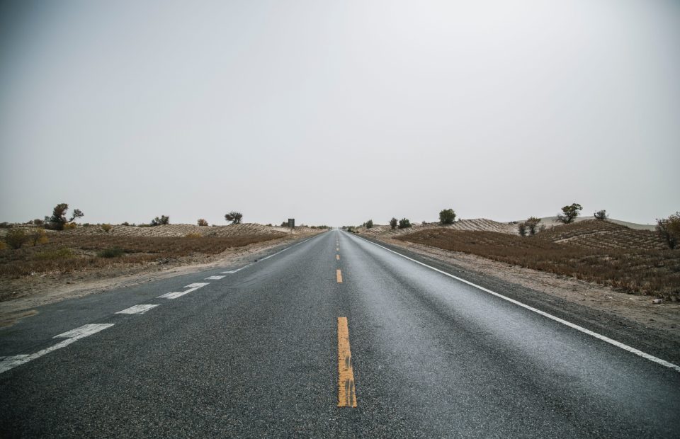 An empty straight road extends into the horizon with a mostly clear sky, flanked by arid, sparsely vegetated land on both sides.