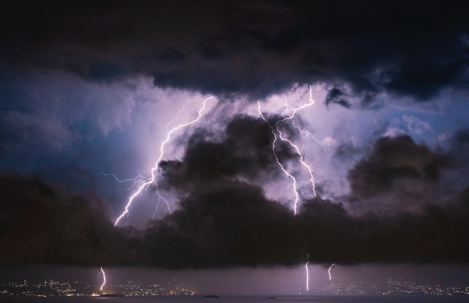 Dark storm clouds with multiple lightning bolts striking over a city at night. Sparse city lights visible along the shoreline.