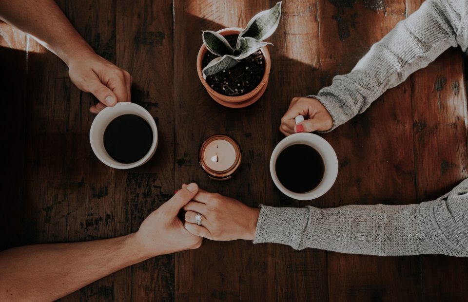 A person with a ring on their finger holds hands with another person, both holding white mugs of coffee over a wooden table, with a small potted plant and a lit candle in the center.