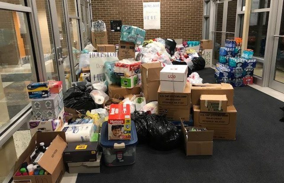 Piles of donation items including boxes of diapers, bottled water, toiletries, and various supplies in a glass-enclosed hallway with a brick wall and a sign that says "Donations Here."