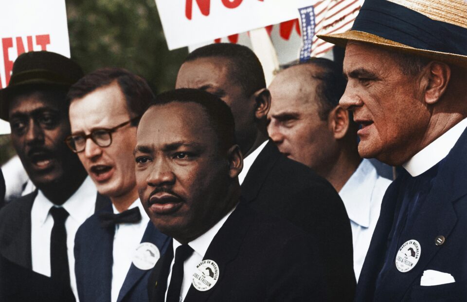 A diverse group of people dressed in formal attire, some with protest signs, participating in a demonstration or rally outdoors.