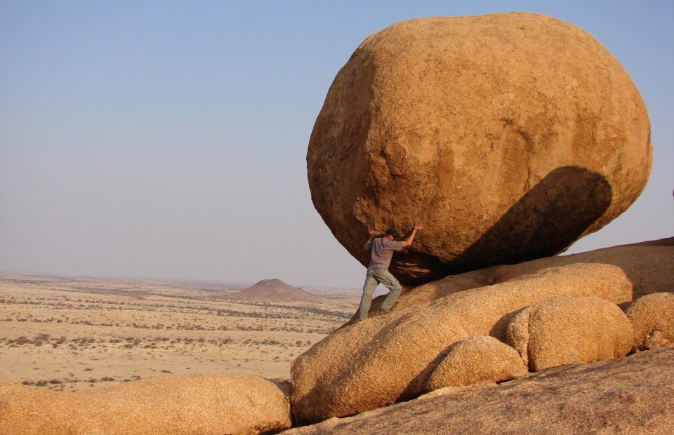 A person in outdoor gear pushes against a large, rounded boulder balanced on smaller rocks in a desert landscape.
