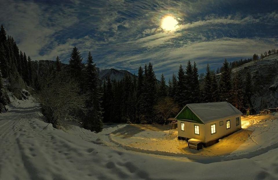 A snow-covered house with lit windows sits beside a wooded mountain landscape under a moonlit sky. The scene is illuminated by the moon and indoor lights.