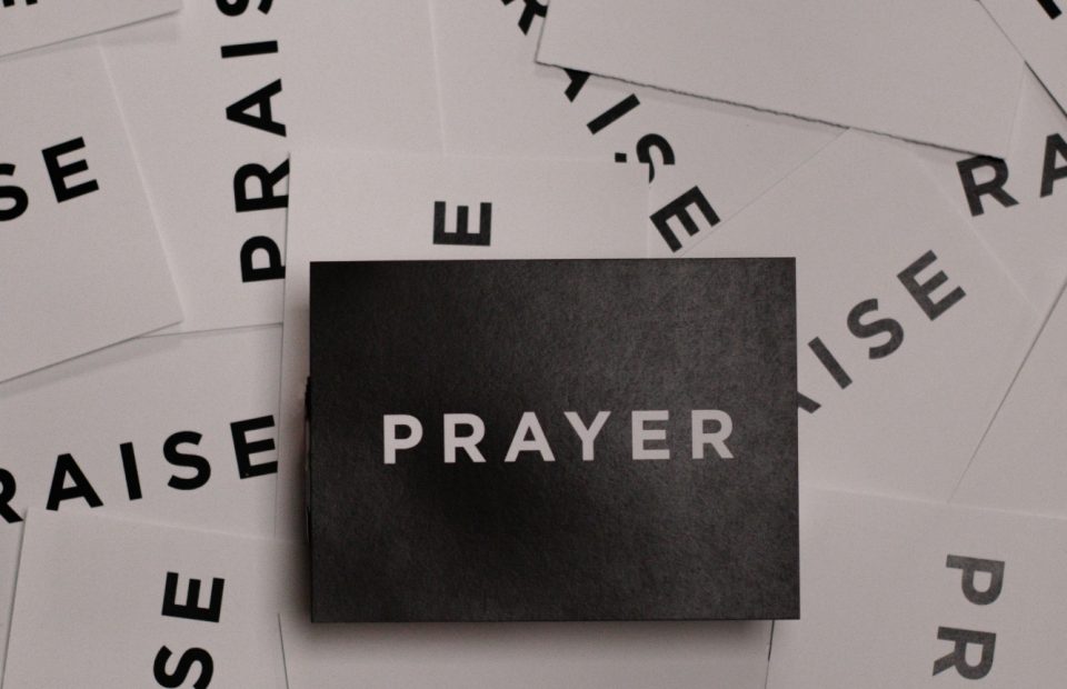 A cluster of white cards with the word "PRAISE" in bold black letters and a black card with "PRAYER" in white letters at the center.