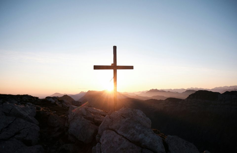 A large wooden cross stands on rocky terrain at sunset, with mountains in the background and the sun partially visible behind the cross.