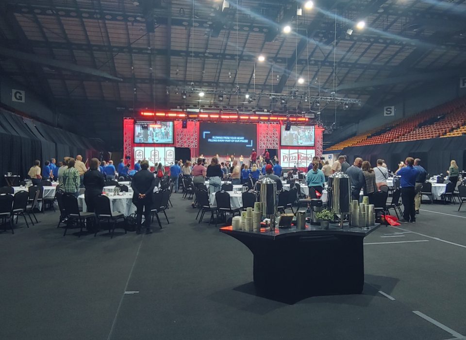 People standing around tables in a large arena singing with a praise and worship band