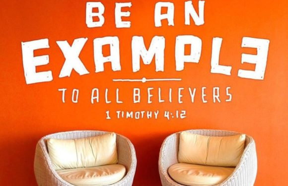 A bright orange wall features a motivational quote about being an example to believers, with two beige cushioned chairs in front of the wall.