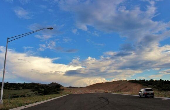 A car driving on an open road with a lamp post on the left and rolling hills in the distance under a blue sky with scattered clouds.