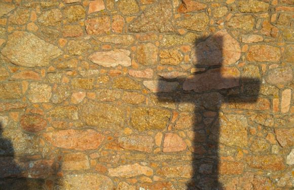 Shadows of a person with arms outstretched are cast on a textured stone wall. The person's shadow appears on a tan and reddish stacked stone surface.