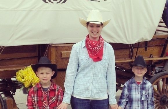 A woman and two boys stand in front of a large white banner with the "Pizza Ranch" logo, dressed in western attire including cowboy hats, plaid shirts, and a red bandana.