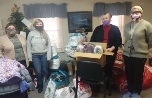 Four women wearing masks stand in a room with holiday decorations, around boxes and bags of donated items, some holding a large open box of supplies.
