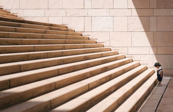 A young child wearing a hat, sunglasses, a striped shirt, and shorts stands at the bottom of large beige stone steps with a stone wall in the background.