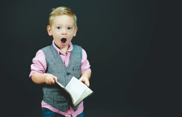 A young boy with a surprised expression, holding an open book, wearing a pink shirt with a gray vest, standing against a dark background.