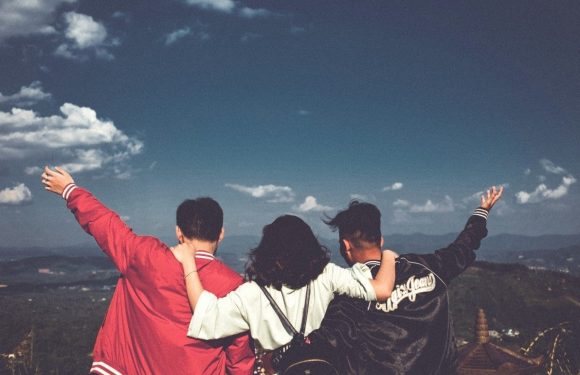Three friends with their arms around each other, facing away, with a scenic landscape and cloudy sky in the background.
